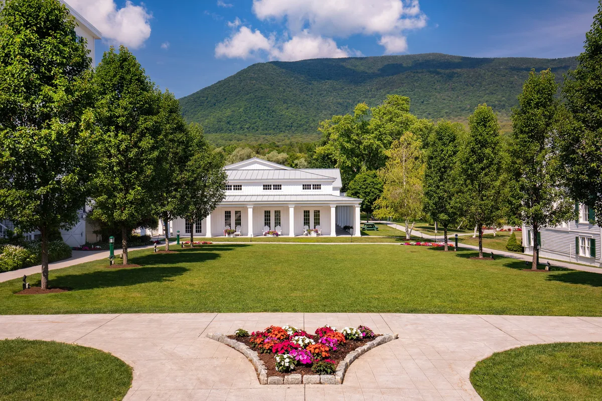 The exterior of the main resort building is shown, framed by colorful flower beds and lush trees, with rolling mountains in the background. The expansive lawn invites outdoor relaxation and exploration, underscored by sunset lighting for a warm, inviting mood.