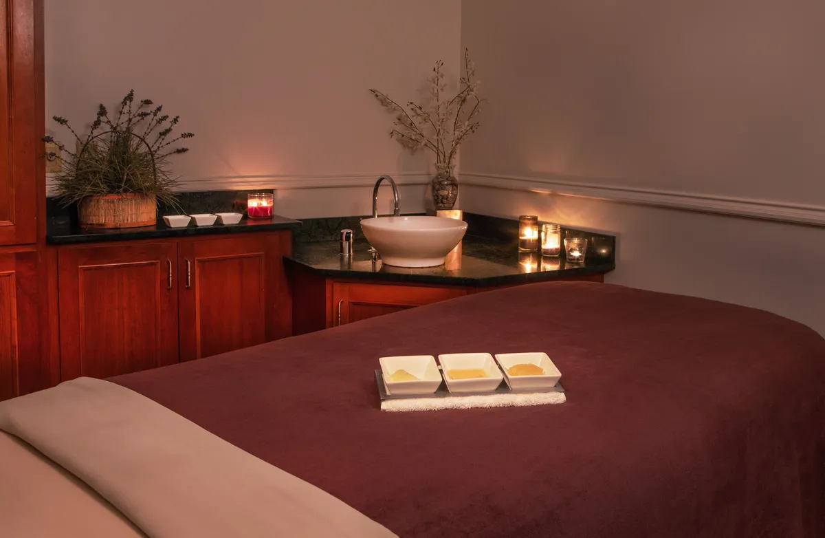 A serene spa room featuring a plush treatment bed with soft linens, bowls of spa treatments ready for use, gentle candlelight, and lush decorative plants. The space exudes calm and invites guests to indulge in rejuvenating spa experiences.
