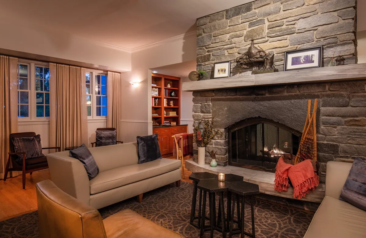 A cozy living room featuring a beautiful stone fireplace, comfortable seating arranged for conversation or relaxation, and bookshelves filled with books and decorative touches. The ambient lighting and plush furnishings create a welcoming space ideal for gathering with friends or family, playing games, or sharing stories by the fire.