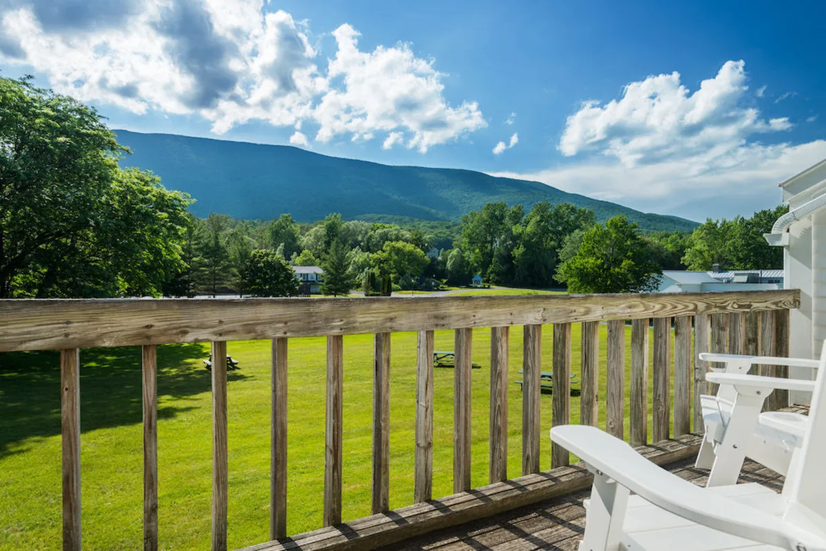 An inviting balcony or patio overlooks lush Vermont greenery and distant mountains. White chairs offer a tranquil spot for sipping coffee at sunrise or enjoying the evening breeze, with a wooden railing framing the natural view.