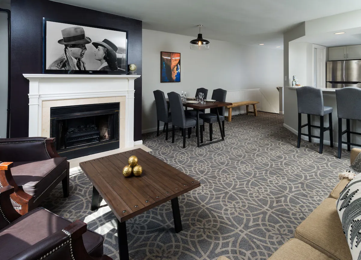 A welcoming, open-plan living and dining area is centered around a stone fireplace, with comfortable sofas arranged for conversation or relaxing. A stylish dining table sits nearby, complemented by tasteful artwork on the walls. Large windows fill the space with natural light, enhancing the cozy, social atmosphere.