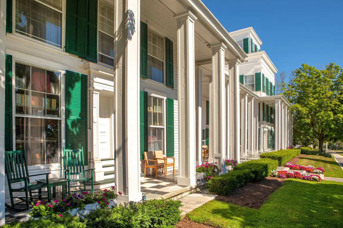 A welcoming front porch with green shutters, classic rocking chairs, and vibrant flower beds offers a serene spot for relaxing or gathering. The inviting columns and well-tended garden evoke a sense of homey comfort and togetherness.