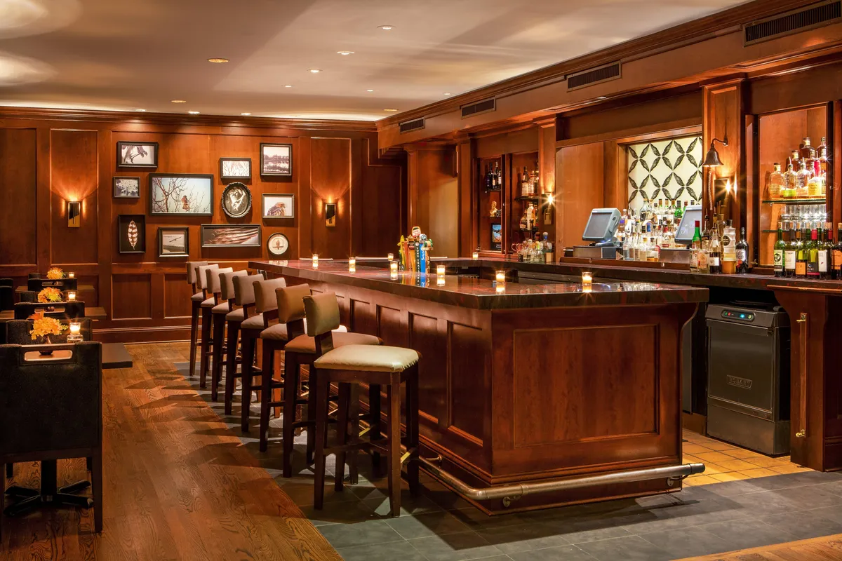 A sophisticated bar area is presented, with high stools, an elegant bar counter, framed artwork, and a display of bottled spirits. The setting is ideal for cocktail tastings, celebratory toasts, or casual mingling before heading out to local hotspots.