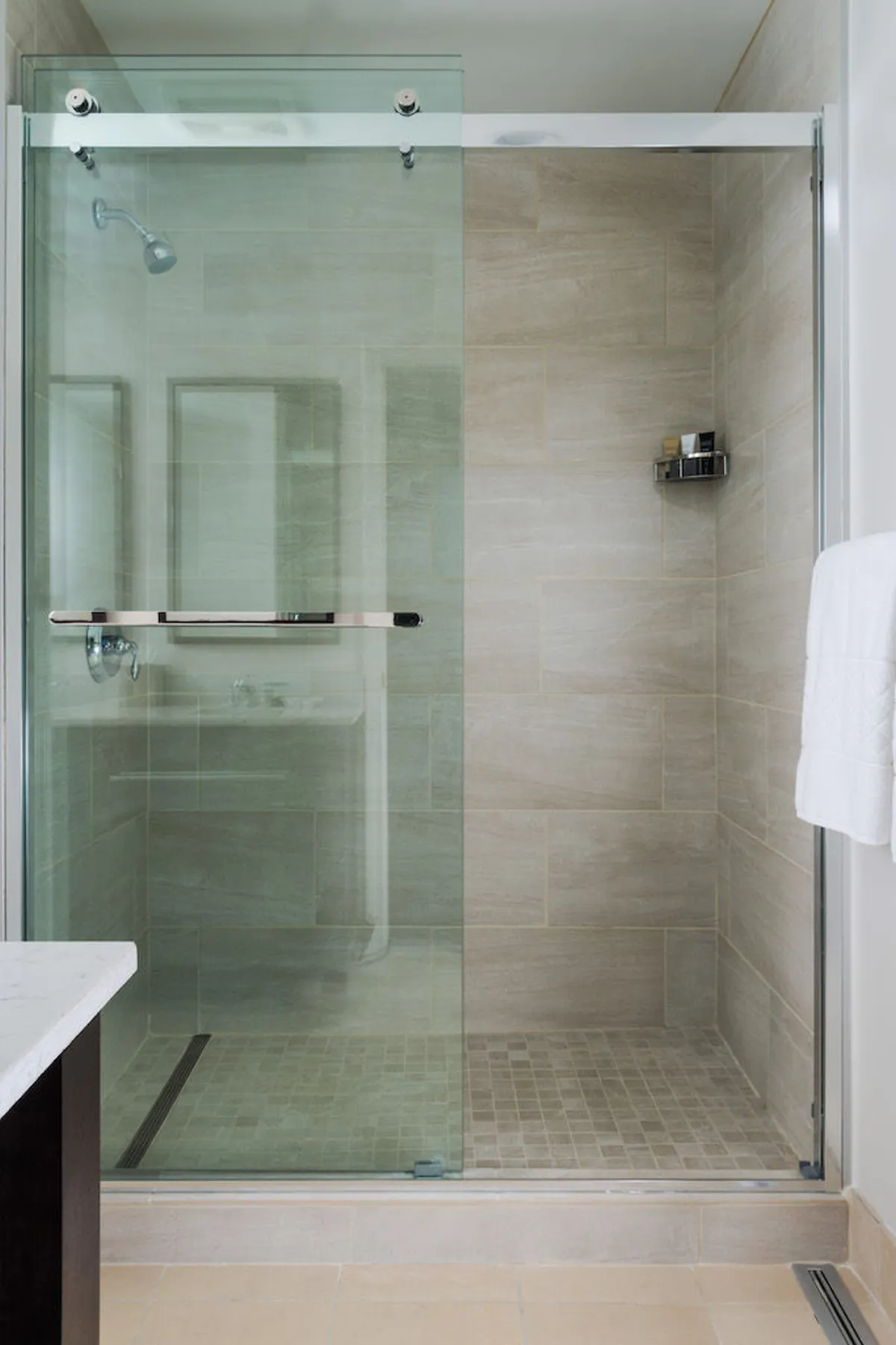 A contemporary bathroom showcasing a glass shower door, textured tiles, a minimalist shelf with quality toiletries, and a plush white towel. The design is modern and evokes a spa-like feel.