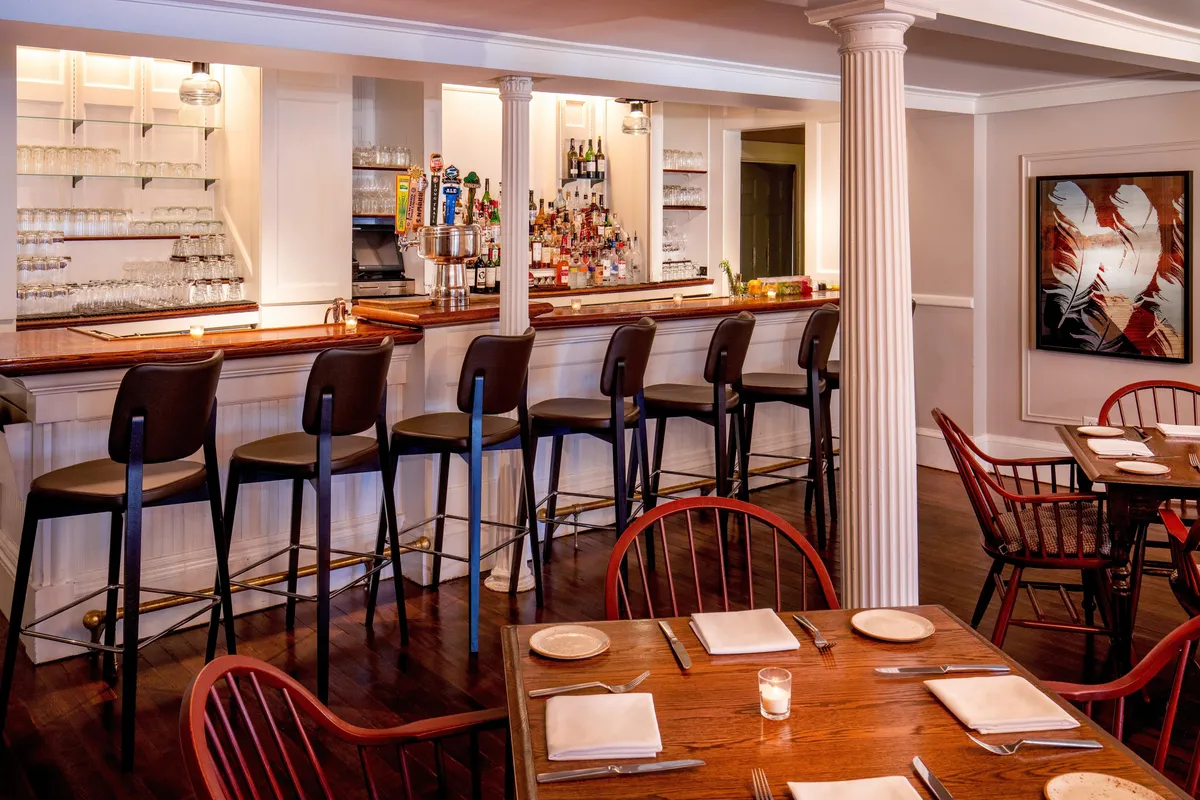 This image showcases a lively interior scene featuring a bar counter with stools, adjacent dining tables, and vibrant artwork on the walls. The arrangement invites guests to gather, enjoy drinks, savor meals, and engage in conversation, echoing the spirit of group getaways and socializing.