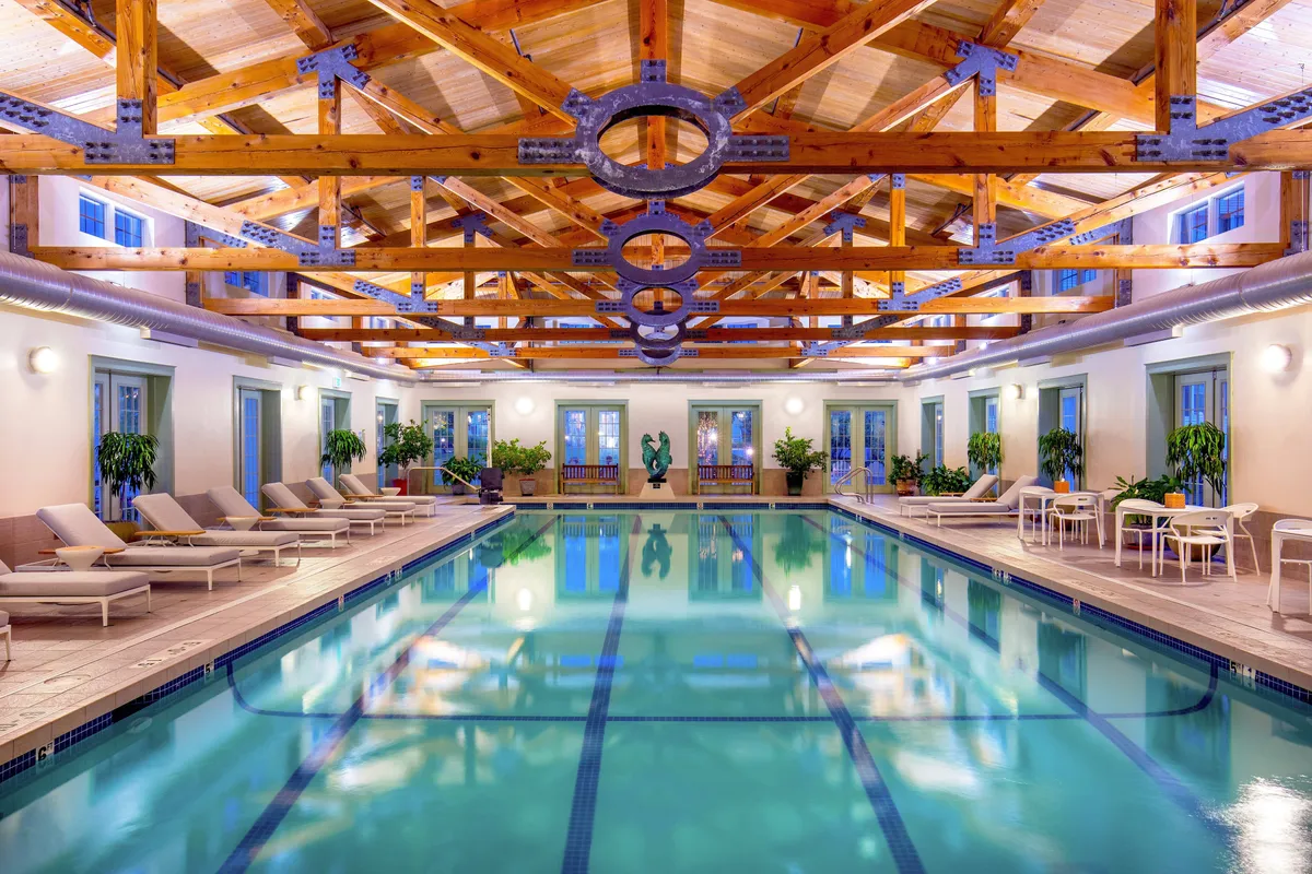 An inviting indoor swimming pool area with elegant wooden beams, plush lounge chairs, lush decorative plants, and a striking sculpture. The tranquil atmosphere is perfect for both swimming and relaxing, with ample space for socializing or enjoying refreshments poolside.