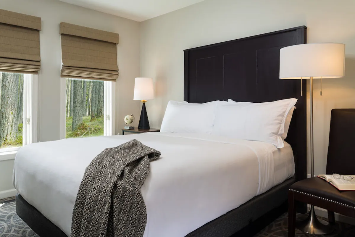 A bedroom featuring a large bed with crisp white linens, a decorative throw blanket, a table lamp on the nightstand, and soft natural light streaming in through the windows. The space exudes a serene and comfortable atmosphere ideal for relaxation.