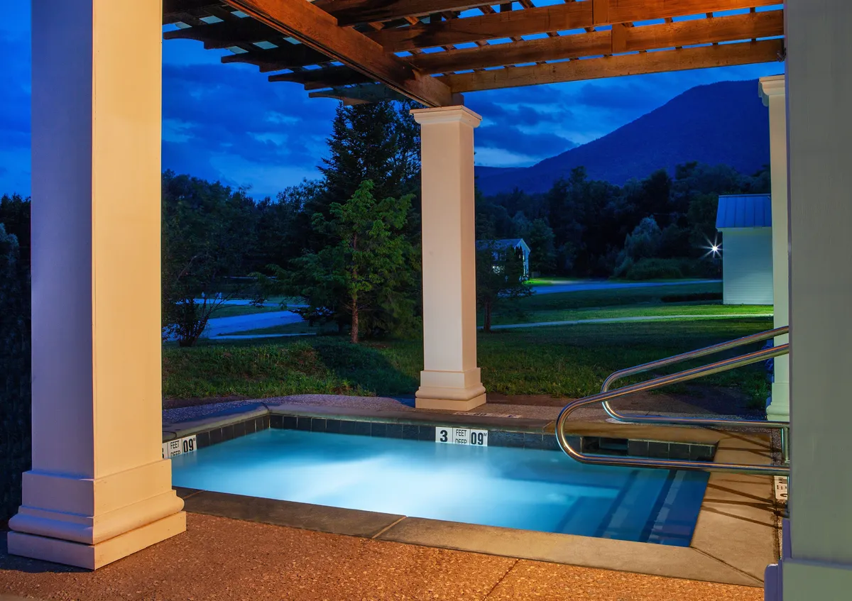 The image captures an illuminated outdoor pool beneath a pergola structure with a stunning mountain view in the background. The tranquil setting features inviting pool waters, warm lighting, and open skies—suggesting both daytime relaxation and evening stargazing opportunities.